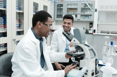 two males, high school student Jordan McDonald, and a medical student Kowshik Sen, sitting on stools in a lab smiling and talking while the older one teaches the younger one how to use a microscope that is sitting on a white counter in the lab 