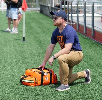 An alumnus of TouroCOM Harlem crouches on a sports field with medical equipment.