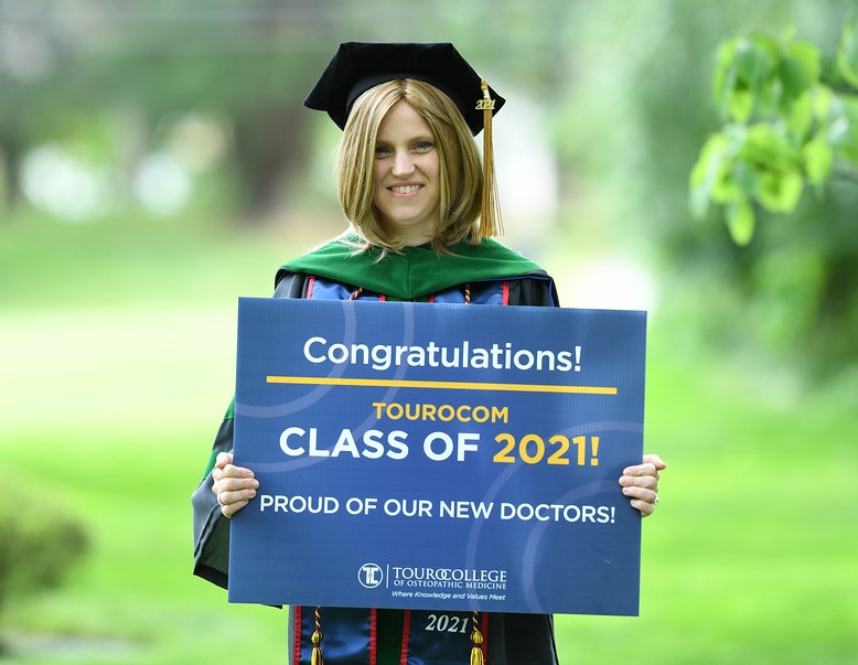 Alexandra Friedman in cap and gown holding a lawn sign that says "Congratulations! TouroCOM Class of 2021! Proud of our new graduates"