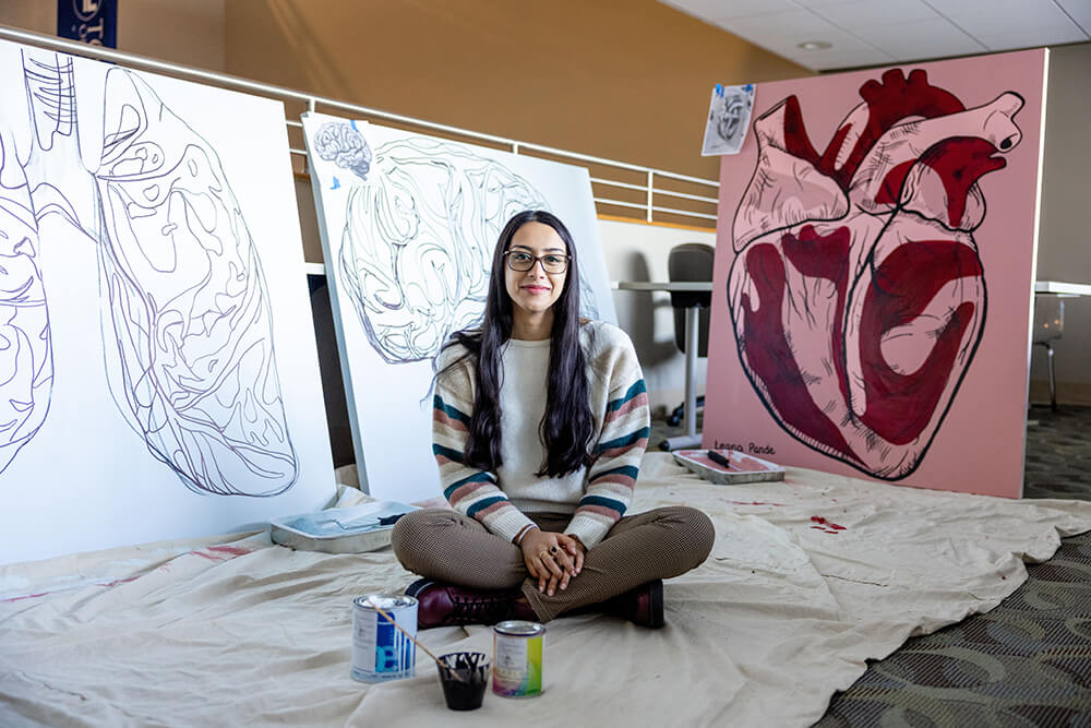 Medical student Leana Pande sitting cross-legged on floor surrounded by large canvases she is painting of human organs. 