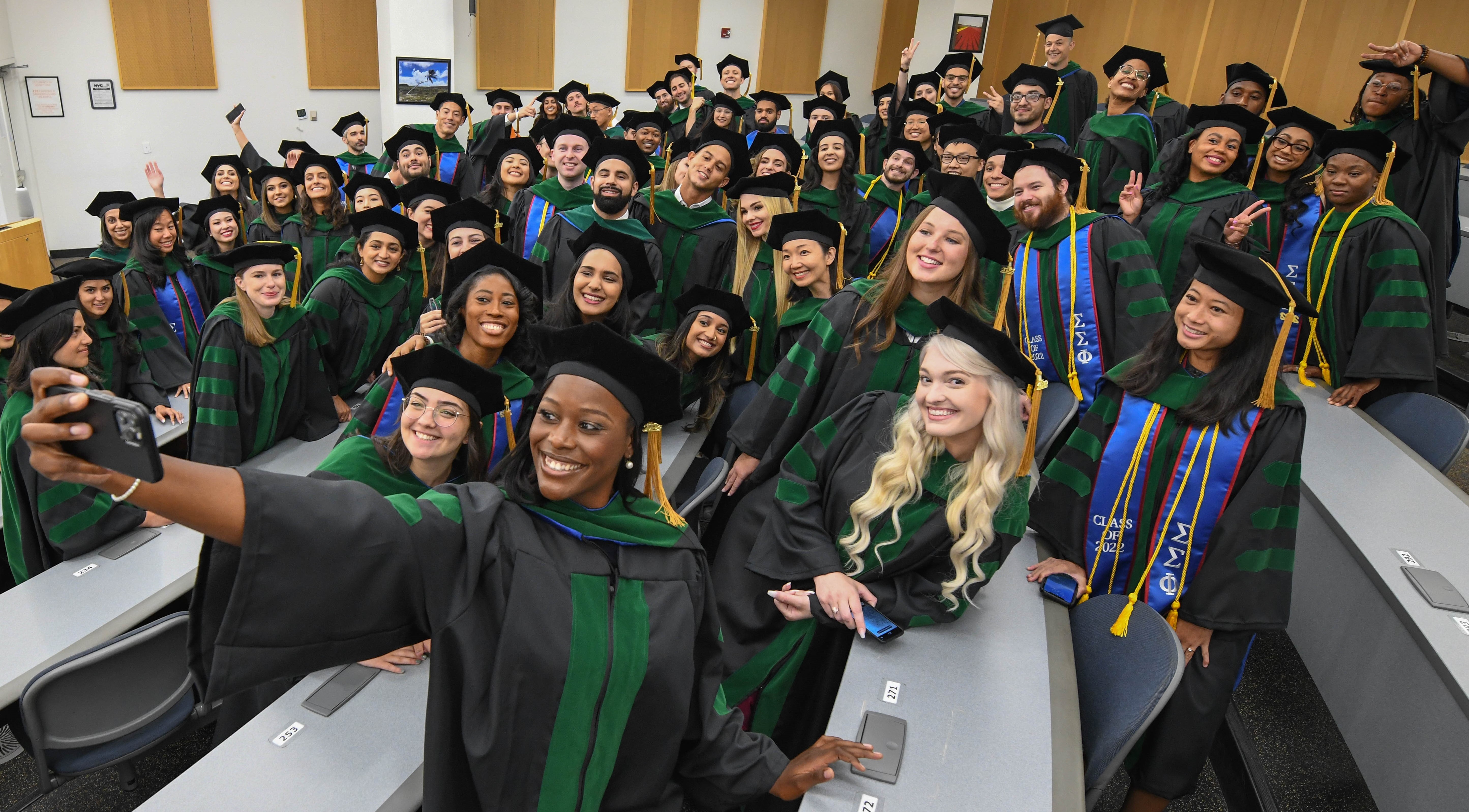 graduates standing in lecture hall taking a group selfie in caps and gowns
