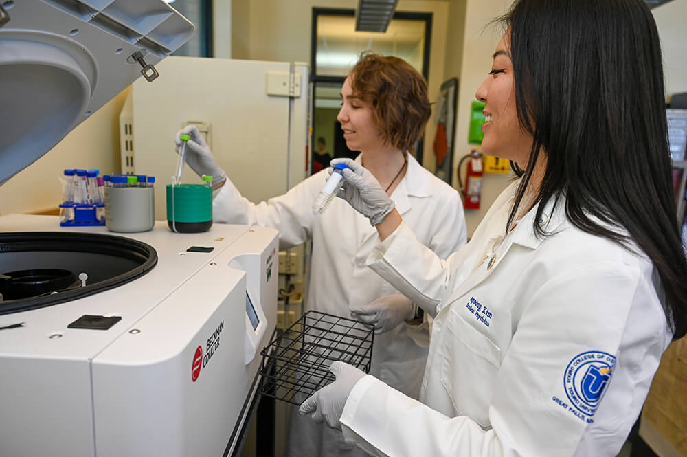 Student physician Jenny Kim smiling while putting test tube into centrifuge in lab with fellow female student