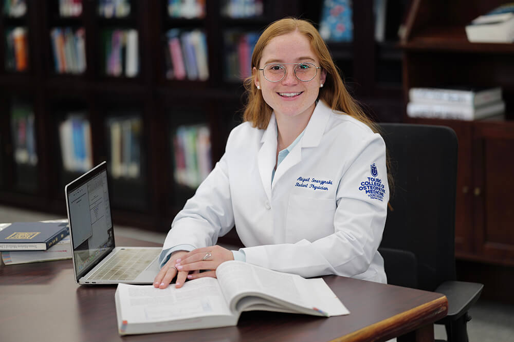 Female student, with long red hair and glasses, in white coat, sitting at a table with an open textbook in library