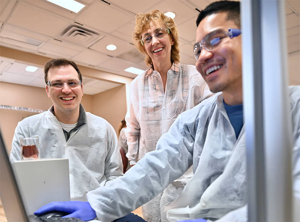 Dr Hanna Wollocko, center, in lab with 2 male students seated in front of her, in lab, looking at computer screen, which is unseen